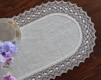 May include: A white oval linen table runner with a delicate lace trim. The lace is a light gray color and has a floral pattern. The runner is on a wooden surface.