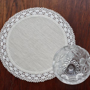 Op de afbeelding: Een ronde, crèmekleurige linnen placemat met een delicate witte kanten rand, gecombineerd met een heldere, geslepen glazen kom. De kom heeft een gedetailleerd, geometrisch patroon. Op een donkerhouten oppervlak.