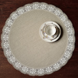 Può includere: Un tovaglietta di pizzo bianco con un delicato motivo floreale si trova su una superficie di legno scuro. Una tazza da tè bianca e un piattino con un design floreale sono posizionati al centro della tovaglietta.