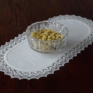 Può includere: Una ciotola di vetro trasparente e sfaccettato piena di fiori secchi è appoggiata su una tovaglietta ovale bianca con un delicato bordo in pizzo. La tovaglietta è posizionata su una superficie di legno scuro.
