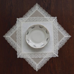 May include: A white porcelain plate with a floral design sits on a white linen square with a lace border. The square is on a dark brown wooden surface.