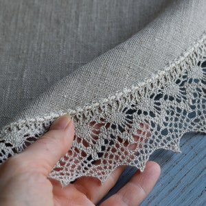 May include: A round, beige linen tablecloth with a delicate lace trim. The lace is a light beige color and has a scalloped edge.