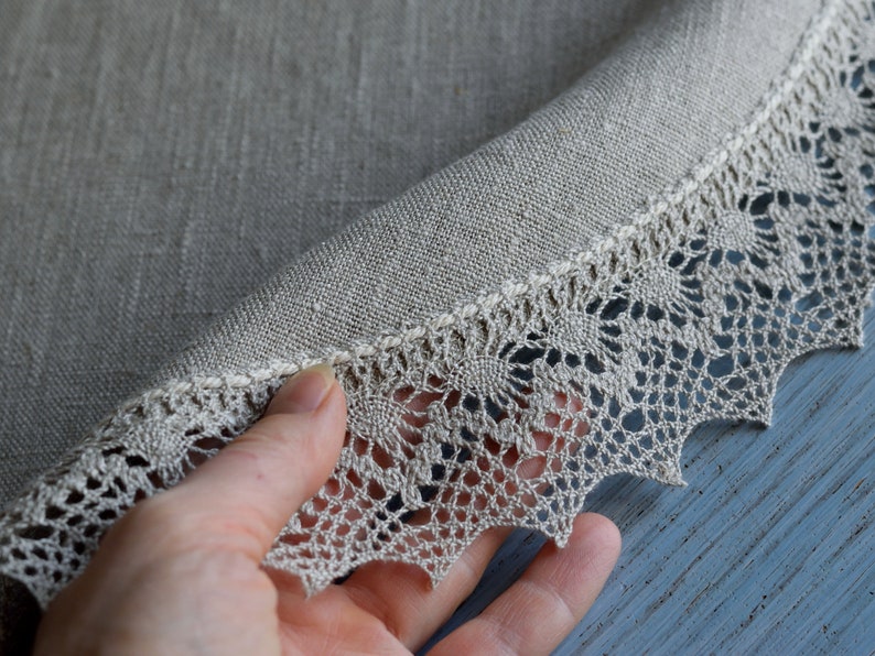 May include: A round, beige linen tablecloth with a delicate lace trim. The lace is a light beige color and has a scalloped edge.