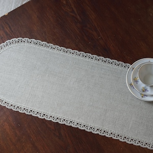 Può includere: Un runner da tavolo in lino grigio chiaro con un bordo in pizzo bianco. Il runner è su un tavolo in legno marrone scuro con una tazza da tè bianca e un piattino con un motivo floreale.