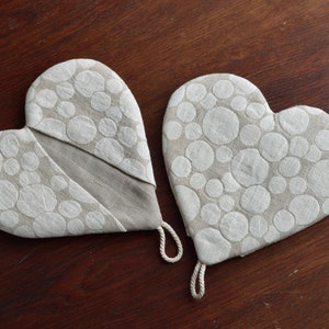 Pode incluir: Duas luvas de cozinha em forma de coração feitas de tecido de linho bege com um padrão de círculos brancos. Cada luva tem uma pequena alça de corda presa no canto inferior para pendurar. O design inclui uma abertura diagonal.
