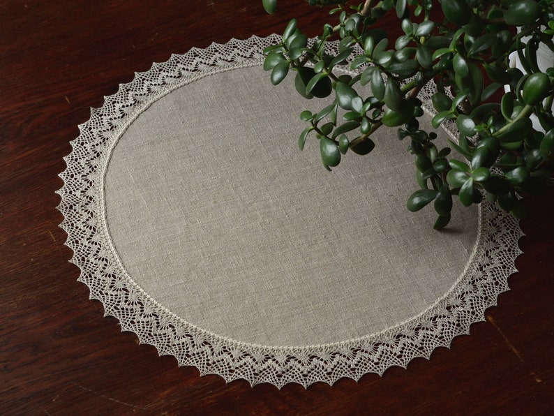 May include: A round, beige linen placemat with a delicate white lace trim. The placemat is on a dark wood table.
