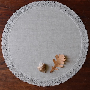 May include: A white round linen tablecloth with a delicate lace border. A brown leaf and a seashell are placed on the tablecloth.