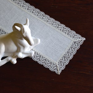 May include: A white linen table runner with a detailed lace trim, set on a dark wooden surface. A white ceramic bull sculpture is placed on the runner, adding a touch of elegance to the scene.