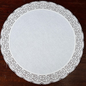 Puede incluir: Un tapete redondo de lino blanco con un delicado borde de encaje floral. El tapete está centrado en una superficie de madera marrón oscuro, mostrando su intrincado diseño y textura. Esta pieza decorativa es ideal para añadir un toque de elegancia a cualquier mesa.