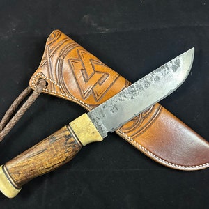 May include: A brown leather knife sheath with a braided strap and a decorative design. A knife with a wooden handle and a metal blade is inside the sheath.