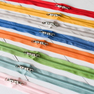 Pode incluir: Conjunto de fitas de zíper coloridas com deslizadores prateados. Os zíperes estão dispostos em uma fila, com cores que vão do vermelho ao rosa.