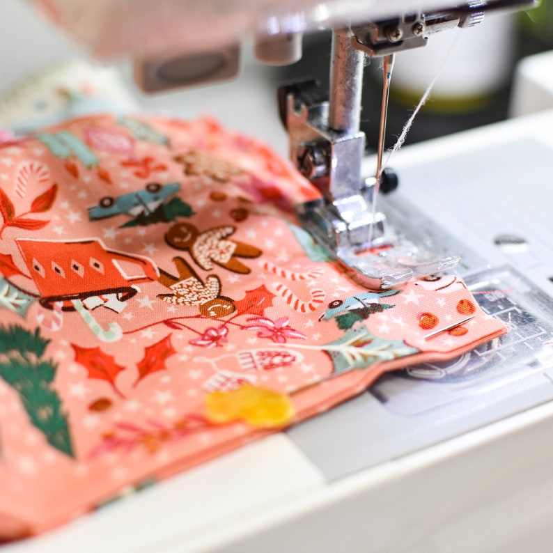 May include: A close-up of a sewing machine needle stitching a pink fabric with a festive pattern of gingerbread people, candy canes, and stars. The fabric is being sewn together to create a handmade item.