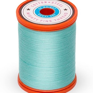 May include: A spool of light blue cotton thread with a white label that reads "Cotton Steel Mighty Fine Thread 690 Yards 50 Weight".