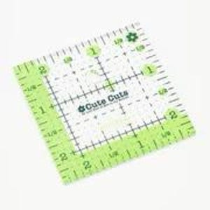 May include: A green and white plastic quilting ruler with markings in inches. The ruler is square and has a grid pattern with markings for 1/8, 1/4, 1/2, and 1 inch. The ruler has the text "Cute Cuts" printed on it.