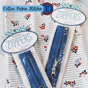 Puede incluir: Dos cremalleras azules en embalaje transparente se muestran sobre un fondo de tela floral. La tela tiene una base blanca con rayas grises y pequeñas flores rojas y azules. Las cremalleras tienen tiradores de metal plateado y están etiquetadas como "Happy Zippers".