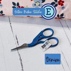 May include: Blue fabric scissors with a curved blade and a white label that says "Made in China". The scissors are on a white wooden surface. The text "Cotton Pickin Stitchin E" is visible in the background.