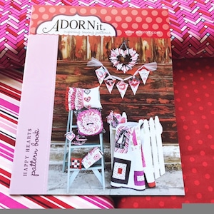 May include: A pink and red book with the title "Happy Hearts pattern book" and the text "AdornIt. Inspiring sewing patterns www.adornit.com". The book features a photo of a rustic wooden background with a white picket fence, a ladder, and various handmade items, including a heart-shaped wreath, pillows, and a quilt.