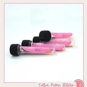 May include: Three clear plastic cylindrical tubes with black screw-on caps, each containing sewing needles. Each tube features a pink label with the text "BUTTERFINGERS #11" and "MILLINERS" printed in white, arranged in a staggered formation.