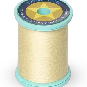 May include: A spool of light beige cotton thread with a blue and yellow label that reads "Cotton + Steel Extra Fancy 50 Weight 680 Yards".