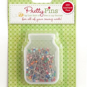 Op de afbeelding: Een doorzichtige plastic pot gevuld met 250 kleurrijke applicatienaalden. De pot is gelabeld "Pretty Pins" door Lori Holt of Bee in my Bonnet. Het label zegt ook "voor al je naaibehoeften!"