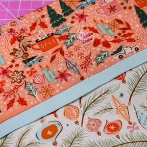 May include: Two pieces of fabric with a Christmas theme. The top fabric is pink with a white background and features a variety of Christmas-themed illustrations, including candy canes, ornaments, and a gingerbread man. The bottom fabric is white with a green background and features a variety of Christmas ornaments.