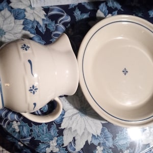 Puede incluir: Juego de jarra y cuenco de color crema con detalles azules. La jarra tiene un asa y un pico, con diseños florales y rayas azules. El cuenco tiene un borde azul y un pequeño diseño floral en el centro. Un juego de menaje clásico.