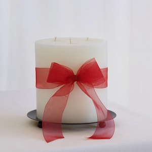 Puede incluir: Una vela de pilar blanca grande con tres mechas, adornada con una cinta roja vibrante atada en un lazo. La vela se encuentra sobre una pequeña placa de metal de color oscuro, creando una pieza decorativa simple pero elegante.