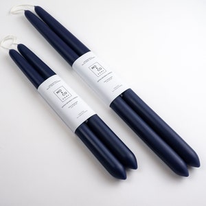 Op de afbeelding: Twee paar marineblauwe dinerkaarsen met witte etiketten. De kaarsen zijn diagonaal op een wit oppervlak geplaatst. Elk paar is verpakt met een wit etiket met zwarte tekst en een logo. De kaarsen hebben verschillende lengtes.