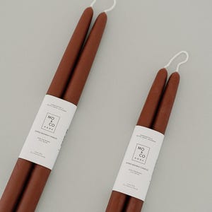 May include: Two brown taper candles with white labels that read "MO & CO HOME" and "DIPPED BEESWAX CANDLES". The candles are made of 100% pure beeswax and have a white string at the top.