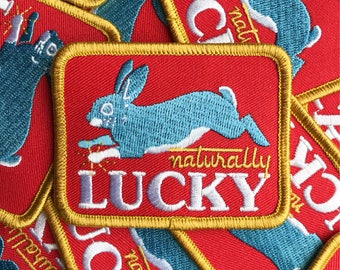 May include: A stack of red embroidered patches with a gold border. The patches feature a blue rabbit with the text "naturally lucky" in white.