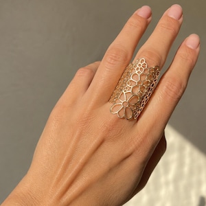 May include: A gold-colored, openwork ring with a floral design. The ring is worn on a hand, and the intricate pattern is visible. The ring's design features a series of connected flower shapes, creating a delicate and elegant appearance.