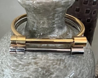 May include: A gold and silver bangle bracelet with a simple, modern design. The bracelet is made of two metal bars connected by a circular band.