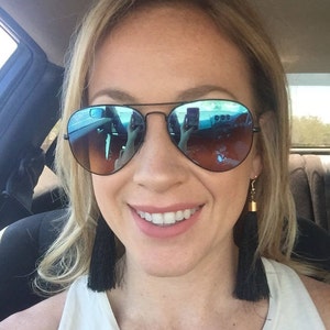 Könnte beinhalten: Eine Frau trägt eine schwarze Aviator-Sonnenbrille mit blauen verspiegelten Gläsern und lange schwarze Quastenohrringe. Sie lächelt und hat blonde Haare.