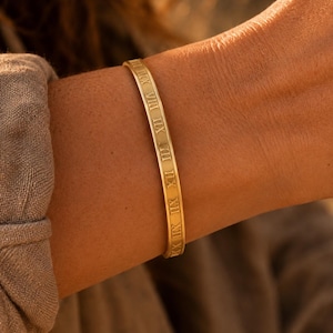May include: A gold-toned metal bangle bracelet featuring a series of engraved Roman numerals along its outer surface. The bracelet has a smooth, polished finish and a slim, uniform band design that encircles the wrist with a consistent width.