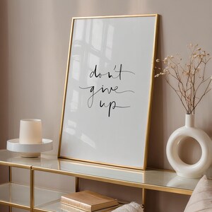 Puede incluir: Un estampado blanco enmarcado con las palabras "don't give up" en escritura negra. El marco dorado se encuentra sobre una consola dorada y de cristal, junto a un jarr&oacute;n blanco con flores secas y un portavelas blanco.