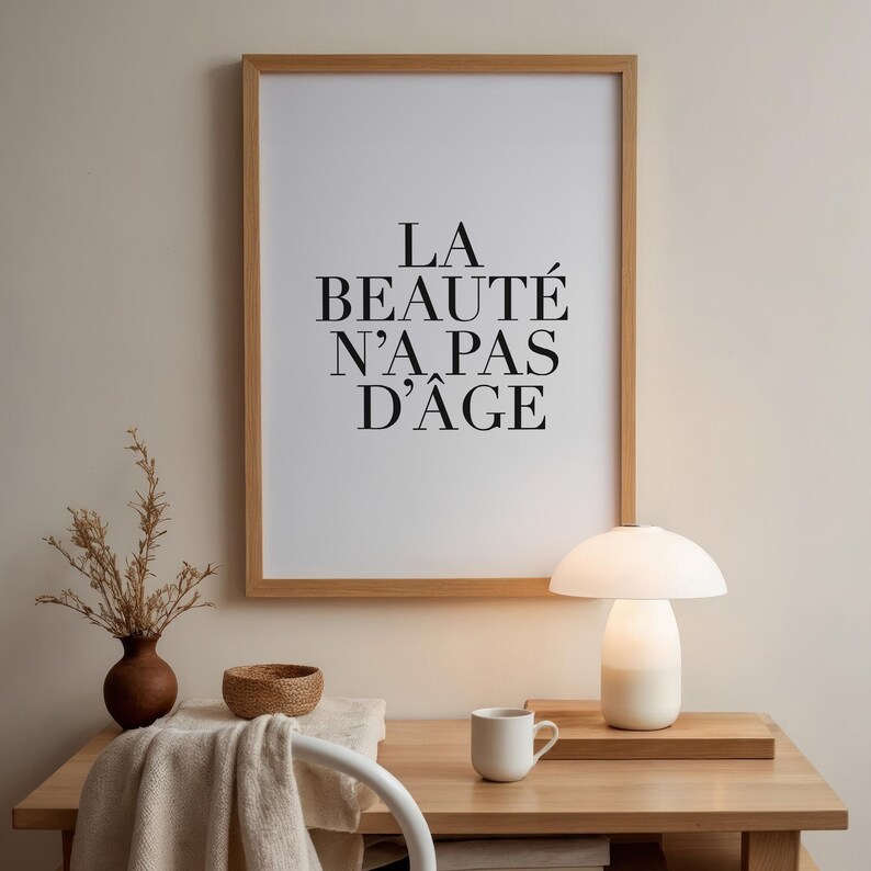 Puede incluir: Una impresi&oacute;n enmarcada con el texto "LA BEAUT&Eacute; N'A PAS D'&Acirc;GE" en letras negras sobre fondo blanco. La impresi&oacute;n est&aacute; sobre un escritorio de madera con una l&aacute;mpara de cer&aacute;mica blanca, un jarr&oacute;n de cer&aacute;mica marr&oacute;n con ramas secas, un cuenco tejido, una taza blanca y una silla blanca.