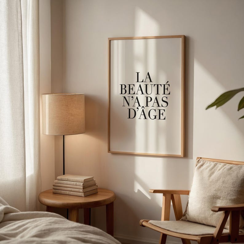 Puede incluir: Una impresi&oacute;n enmarcada con el texto "LA BEAUT&Eacute; N'A PAS D'&Acirc;GE" en letras negras sobre un fondo blanco. La impresi&oacute;n est&aacute; colgada en una pared blanca en una habitaci&oacute;n con una silla de madera, una l&aacute;mpara y una pila de libros sobre una mesa de madera.