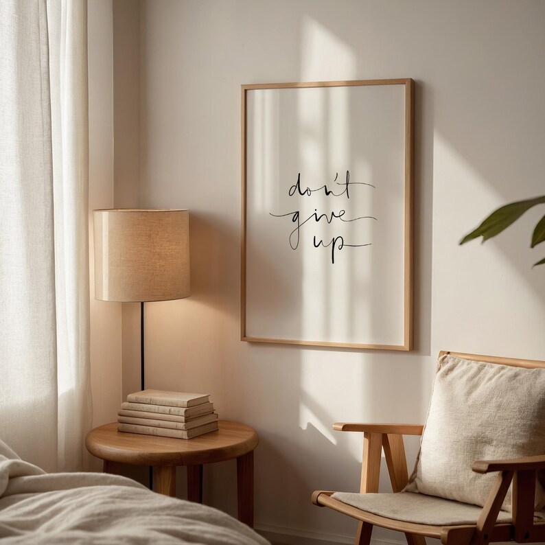 Puede incluir: Un estampado enmarcado con las palabras "don't give up" en escritura negra. El estampado est&aacute; en un marco de madera clara y cuelga de una pared de color neutro. Una peque&ntilde;a mesa auxiliar de madera con libros y una l&aacute;mpara con una pantalla beige est&aacute;n junto al estampado.