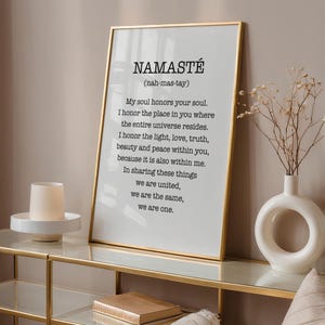 May include: A framed print with a gold frame and a white background. The print features the word "Namaste" in black text with the definition "(nah-mas-tay)" below it. The definition continues with the text: "My soul honors your soul. I honor the place in you where the entire universe resides. I honor the light, love, truth, beauty and peace within you, because it is also within me. In sharing these things we are united, we are the same, we are one."