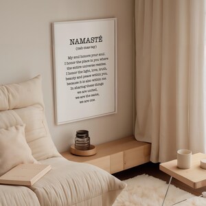 May include: A white framed print with the word "Namaste" in black text. The print includes the definition of the word and its meaning. The print is hanging on a white wall in a room with a couch and a side table.