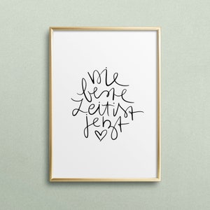 May include: A framed print with a gold frame and a white background. The print features black handwritten text that reads "Die beste Zeit ist jetzt" with a small heart symbol at the bottom.