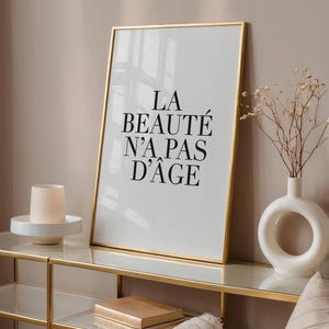 Puede incluir: Una impresi&oacute;n enmarcada en dorado con el texto "LA BEAUT&Eacute; N'A PAS D'&Acirc;GE" en letras negras sobre fondo blanco. La impresi&oacute;n est&aacute; colocada en una repisa dorada con un jarr&oacute;n blanco y un portavelas blanco.