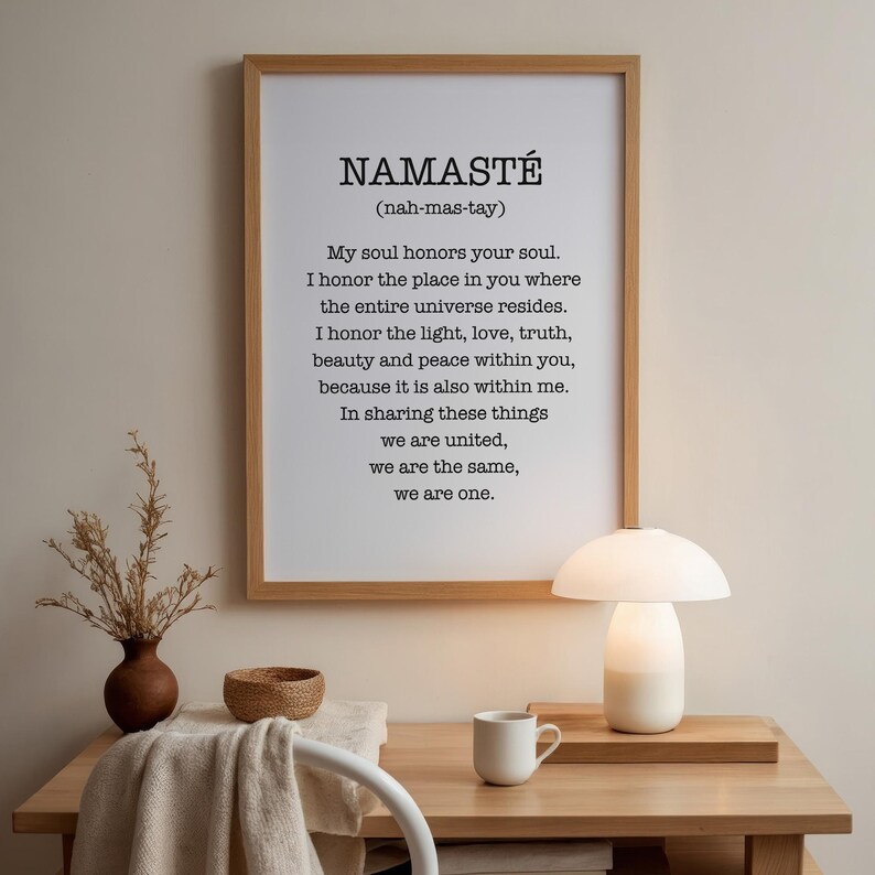 May include: A framed print with the word "Namaste" in black text on a white background. The print includes a definition of the word and its meaning. The print is hanging on a white wall above a wooden table with a white ceramic lamp, a brown ceramic vase with dried branches, a woven bowl, and a white ceramic mug.