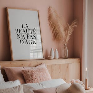 Puede incluir: Una impresi&oacute;n enmarcada con el texto "LA BEAUT&Eacute; N'A PAS D'&Acirc;GE" en letras negras sobre un fondo blanco. La impresi&oacute;n est&aacute; colgada en una pared rosa claro sobre una cabecera de madera con una colcha blanca y almohadas rosas.