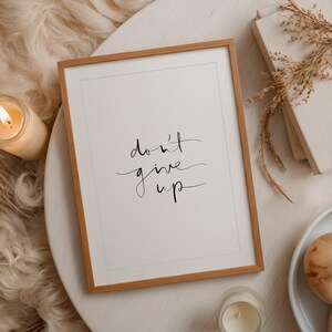 Puede incluir: Una impresi&oacute;n enmarcada con las palabras "don't give up" en elegante escritura negra. El marco es de color marr&oacute;n c&aacute;lido y la impresi&oacute;n est&aacute; sobre un fondo blanco. La imagen est&aacute; sobre una mesa blanca con una vela y un libro.