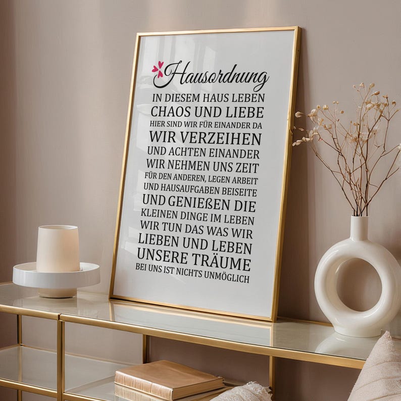 May include: A framed print with a gold frame and a white background. The print features black text that reads "Hausordnung" and a list of rules for living in a home. The text includes phrases like "Chaos und Liebe" and "Wir verzeihen".