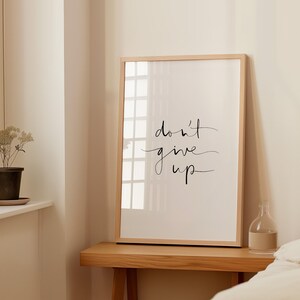 Puede incluir: Un cuadro enmarcado con la frase "don't give up" en elegante escritura cursiva. La obra de arte est&aacute; sobre un fondo blanco y enmarcada en madera clara. La impresi&oacute;n se exhibe sobre una mesa de madera.