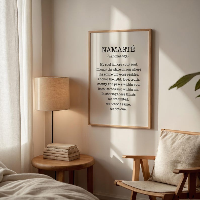 May include: A framed print with a white background and black text. The text reads "NAMASTE (nah-mas-tay)" and continues with a definition of the word. The print is hanging on a white wall in a room with a wooden chair and a lamp.