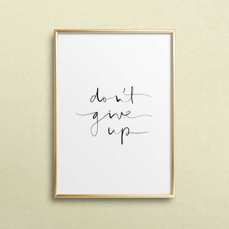 Puede incluir: Un cuadro enmarcado con la frase "don't give up" en elegante escritura negra sobre un fondo blanco. El marco es delgado, de color dorado, y la impresi&oacute;n se muestra contra una pared beige claro.