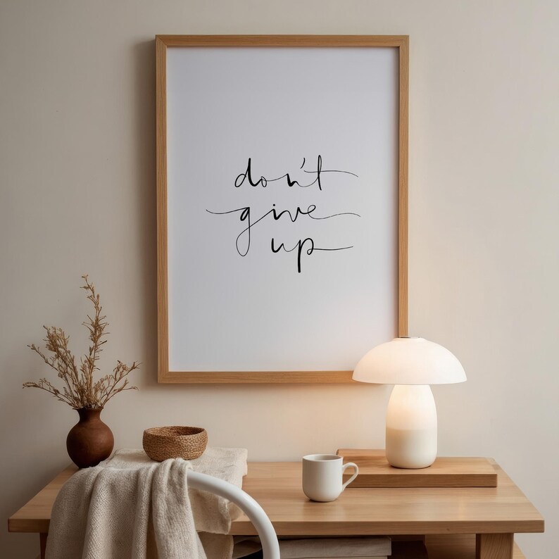 Puede incluir: Un estampado enmarcado con las palabras "don't give up" en una elegante escritura. El marco de madera rodea un fondo blanco. Una peque&ntilde;a l&aacute;mpara blanca, una taza y un jarr&oacute;n con plantas secas est&aacute;n sobre un escritorio de madera.